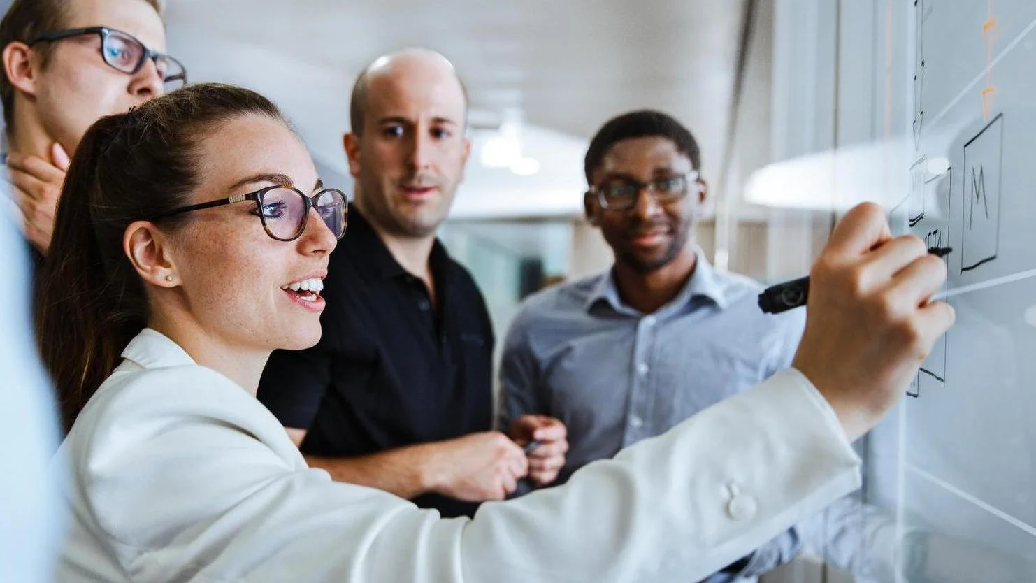 Four professionals stand together in a meeting area while one person writes on a large whiteboard or glass board. The others watch closely as the group discusses ideas or plans. The image shows teamwork, planning, and collaboration at Vector.
