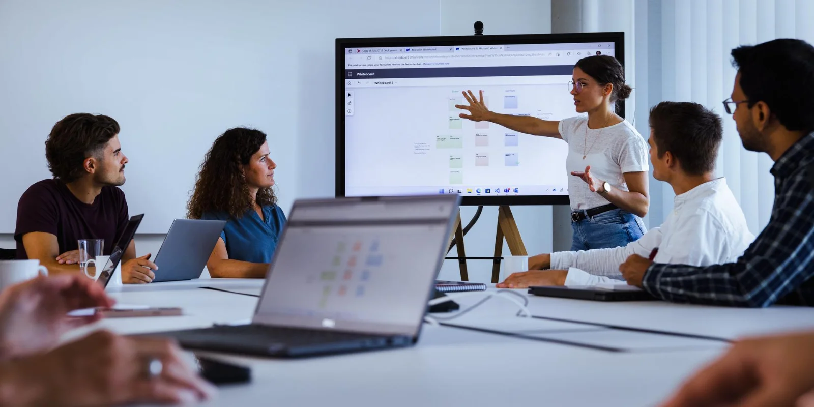 Vector Software Defined Systems helps teams stay in control and scale innovation across the full product lifecycle. A group of professionals sits around a conference table during a team meeting. One person stands near a large screen and points at a presentation while others watch and listen. Several laptops are open on the table, and the setting looks like a modern office meeting room at the software ecosystem company vector.