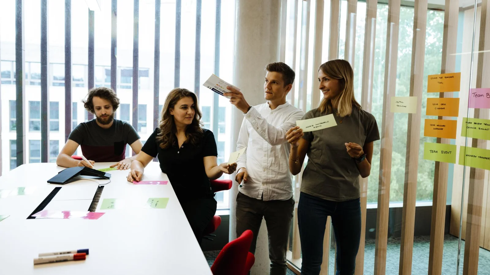 The Vector Foundation puts its resources in research, education, and social cohesion. Four people work together in a bright meeting space at Vector beside a glass wall covered with colorful sticky notes. Two people stand and hold note cards while others sit at a table with papers and markers. The group appears to be discussing ideas during a collaborative workshop or planning session.