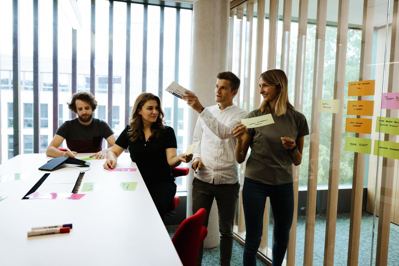 Four people work together in a bright meeting space at Vector beside a glass wall covered with colorful sticky notes. Two people stand and hold note cards while others sit at a table with papers and markers. The group appears to be discussing ideas during a collaborative workshop or planning session.