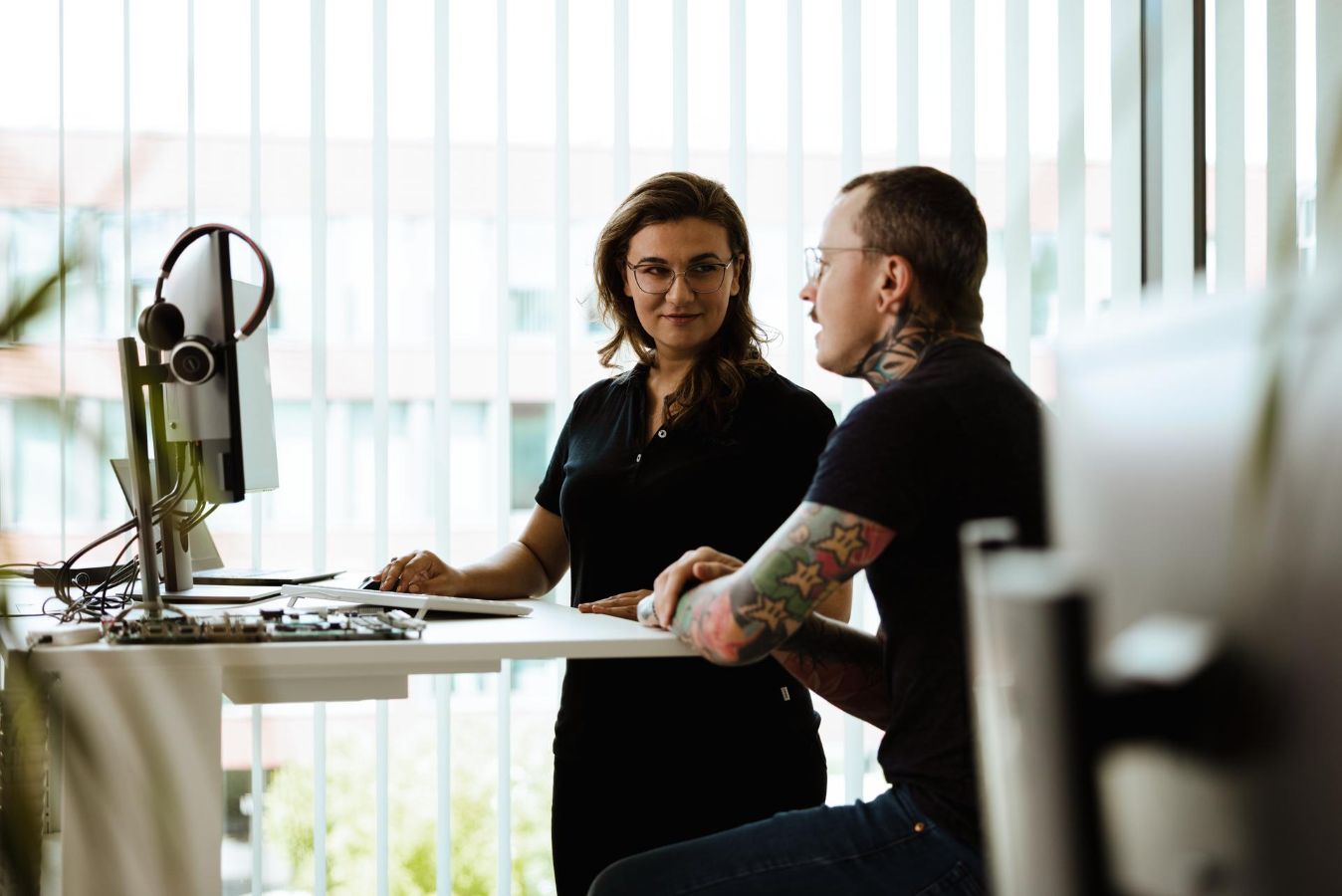 Vector’s whistleblowing system supports safe, confidential reporting and transparent communication. Two Vector employees talk at a workstation in an office. One employee stands beside a desk with a computer and headset, while the other sits and faces them. Large vertical window blinds fill the background.