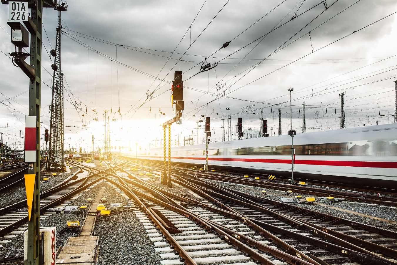 Vector supports evolving rail systems with development and validation workflows that improve long-term operational reliability. Image of multiple railway tracks, overhead power lines, and a fast passenger train moving through a bright rail yard at sunrise.